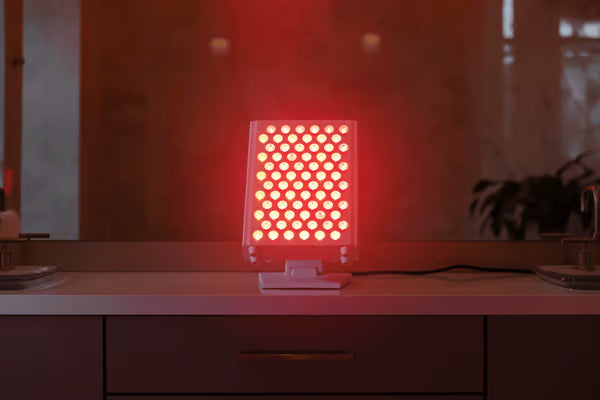 Vital Red LIght. Red Light Therapy. A vertical red light therapy panel on wheels is plugged in and illuminated in a modern living room near a green armchair and a window.